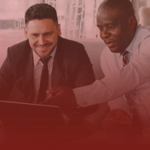 A photograph of two men working in the office of the CFO, looking at a laptop. The man on the left wears a suit and a loose-fitting tie; he is smiling. The man on the right wears a shirt and tie; he is pointing with his hand to the laptop screen, clearing showing the other person some pertinent information.